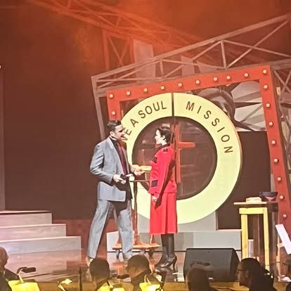 A man in a suit and a woman in a red coat on a stage with a sign that says be a soul mission