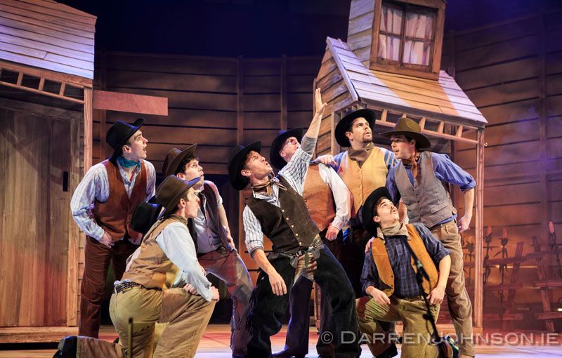 A group of cowboys are standing in front of a wooden building on a stage.
