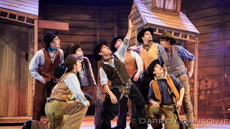 A group of cowboys are standing in front of a wooden building on a stage.