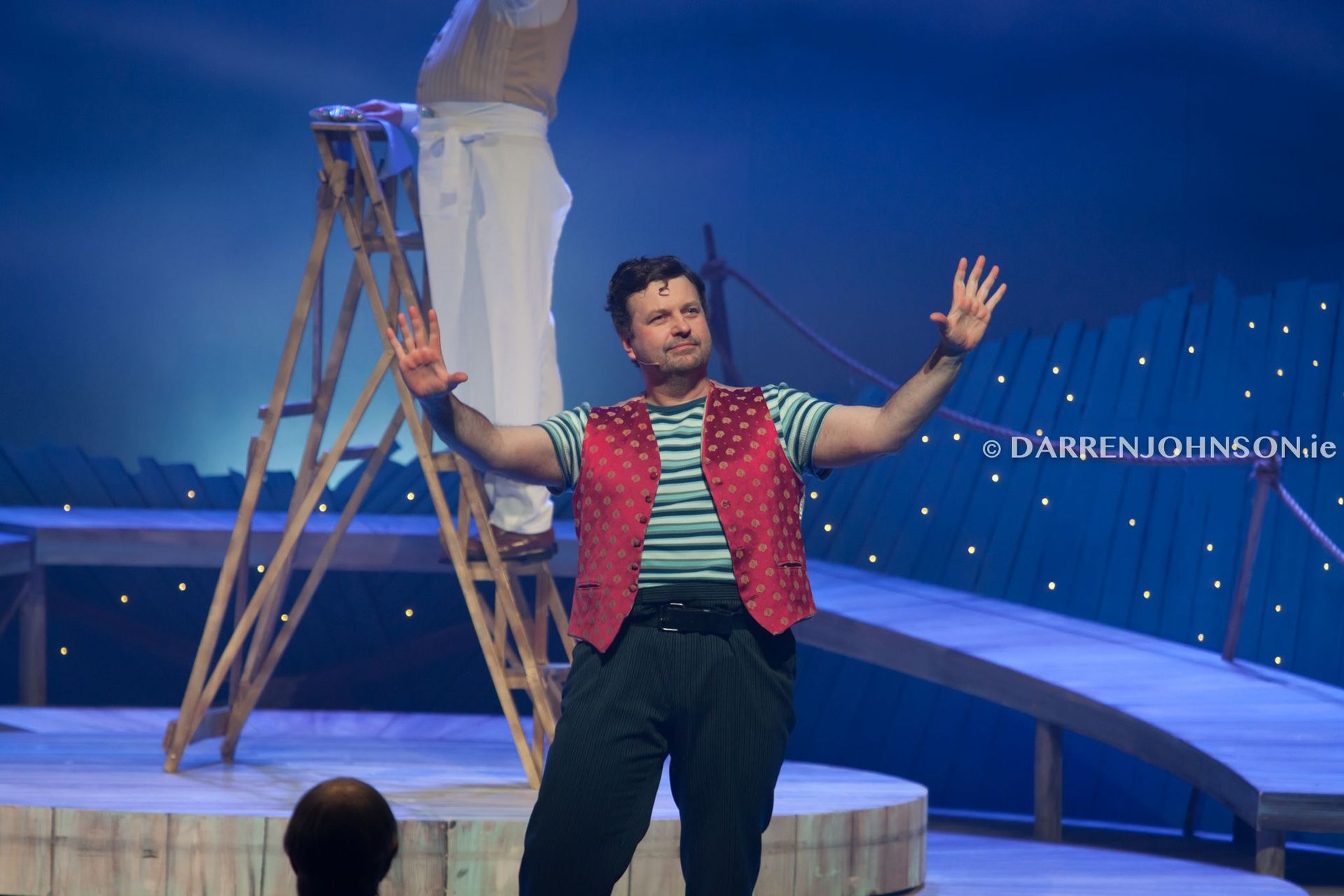 A man in a red vest is standing on a stage with his arms outstretched