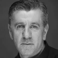 A bald man wearing a black sweater and white shirt smiles for the camera