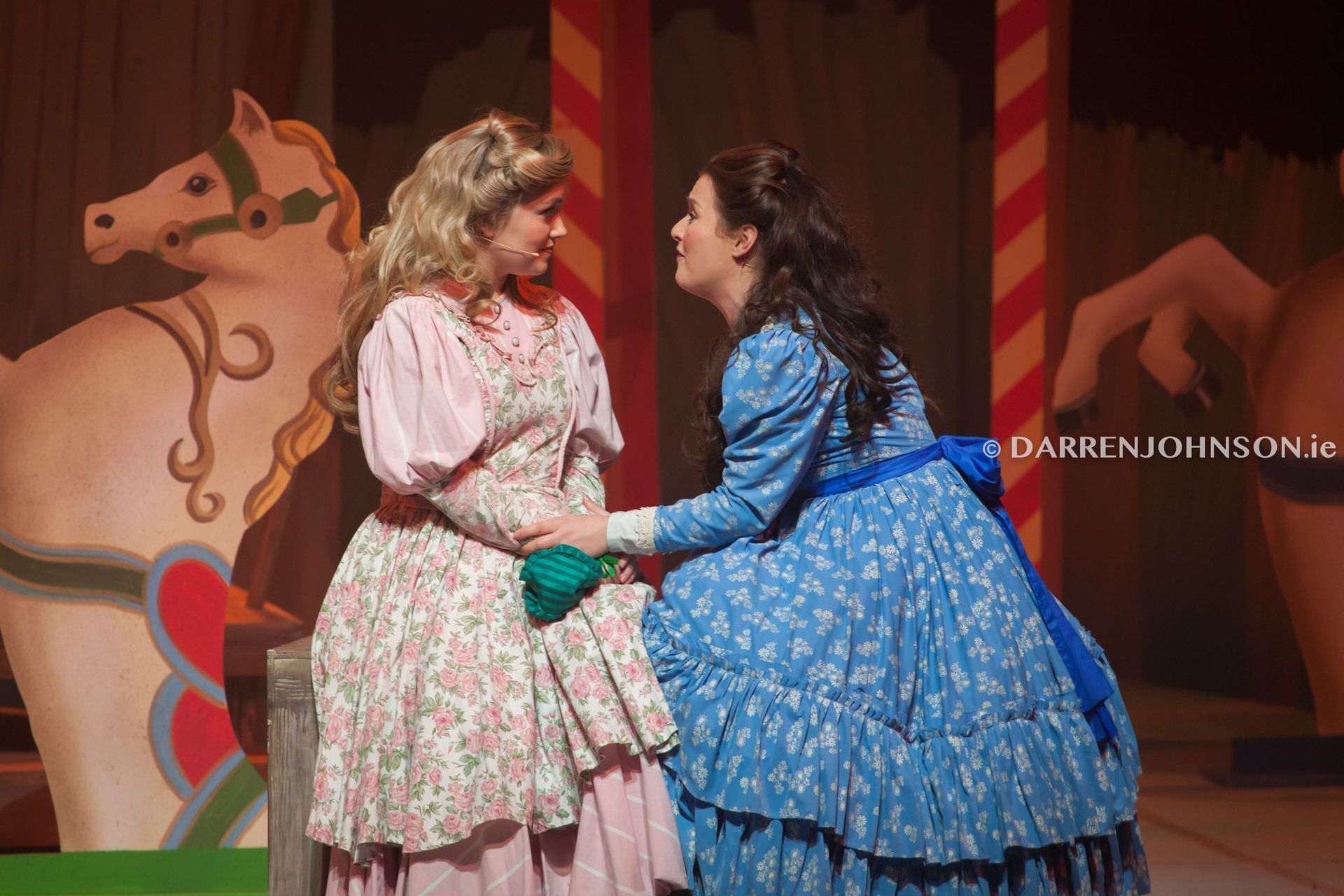 Two women are standing next to each other on a stage in front of a merry go round.