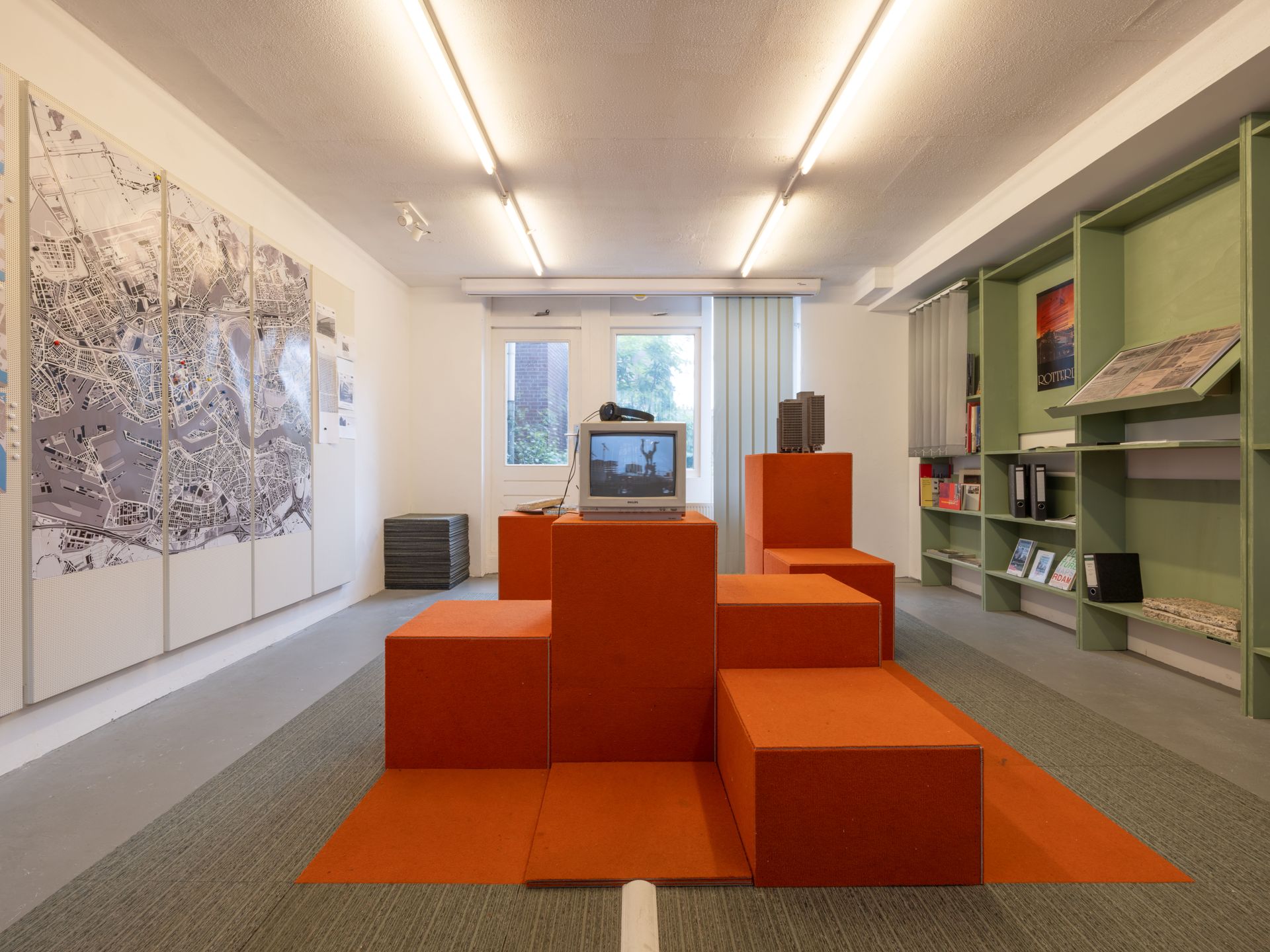 Exhibition landscape made out of carpet tiles, featuring a model and a television, and a map of Rotterdam and an archive.