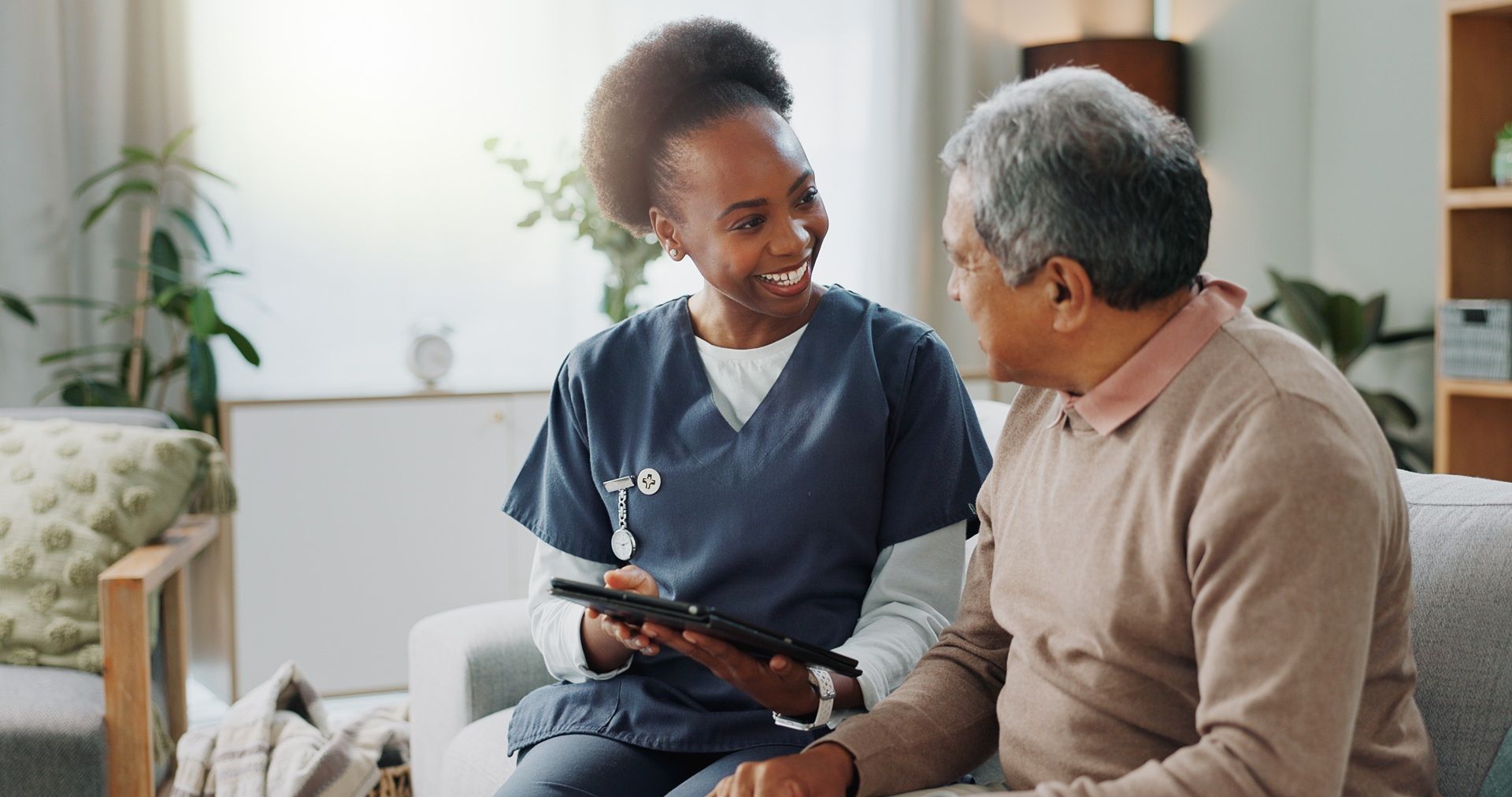 A nurse is sitting on a couch with an elderly man and looking at a tablet.