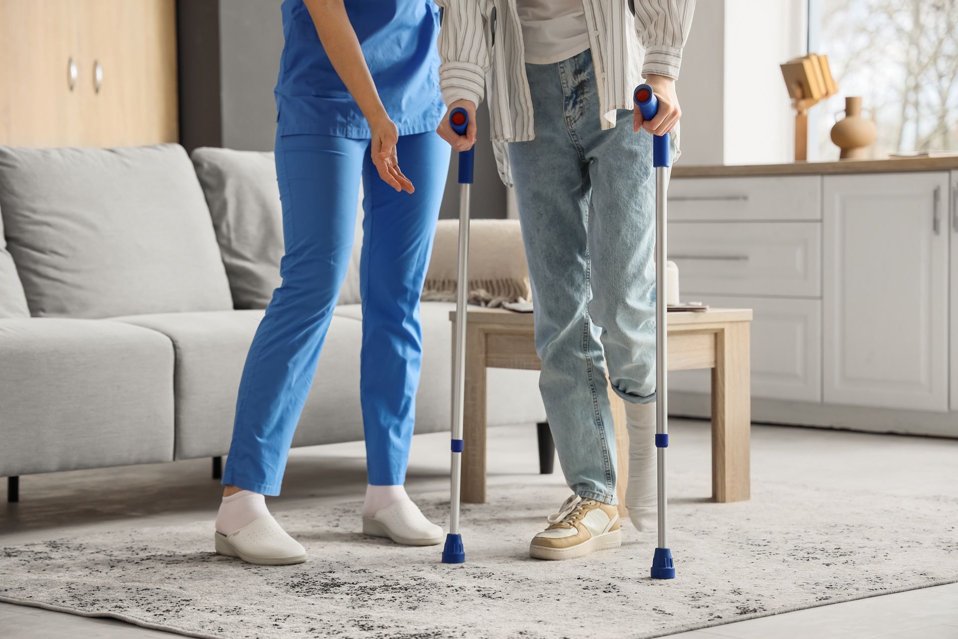 A nurse is helping a man with crutches walk in a living room.