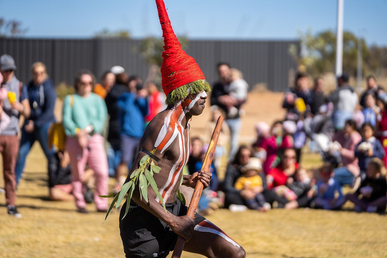 A man with a red hat and a stick is dancing in front of a crowd.