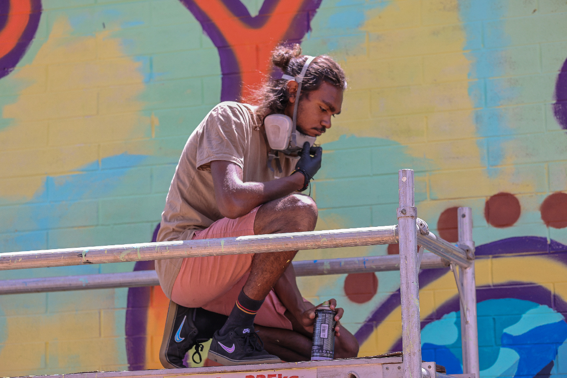 A man is kneeling on a scaffolding in front of a graffiti wall.