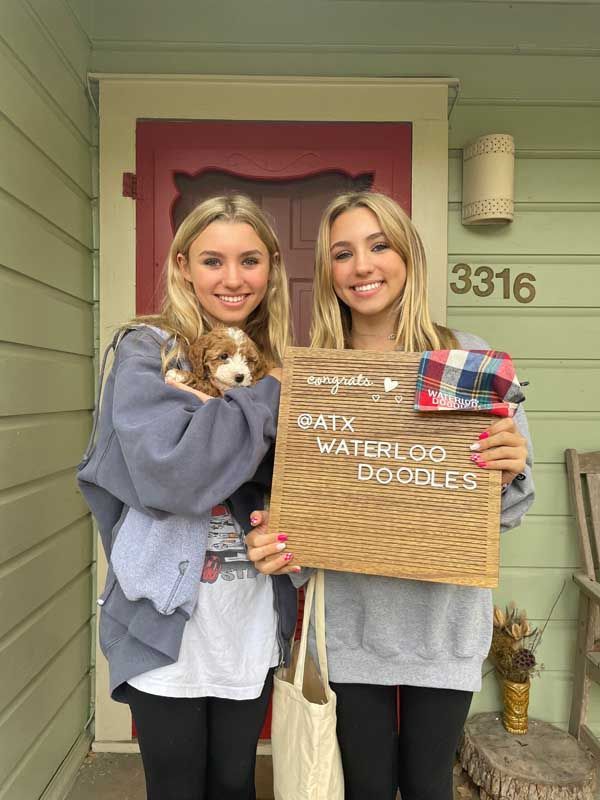 Two women are holding a puppy and a sign in front of a house.