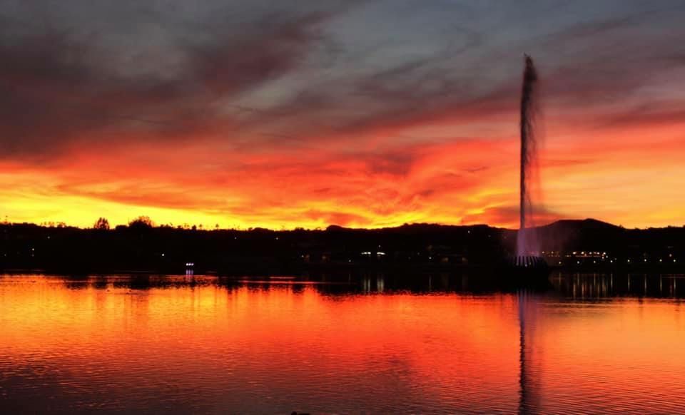Fiery sunset over a lake, silhouetted trees and a fountain. Reds and oranges reflected in the water.