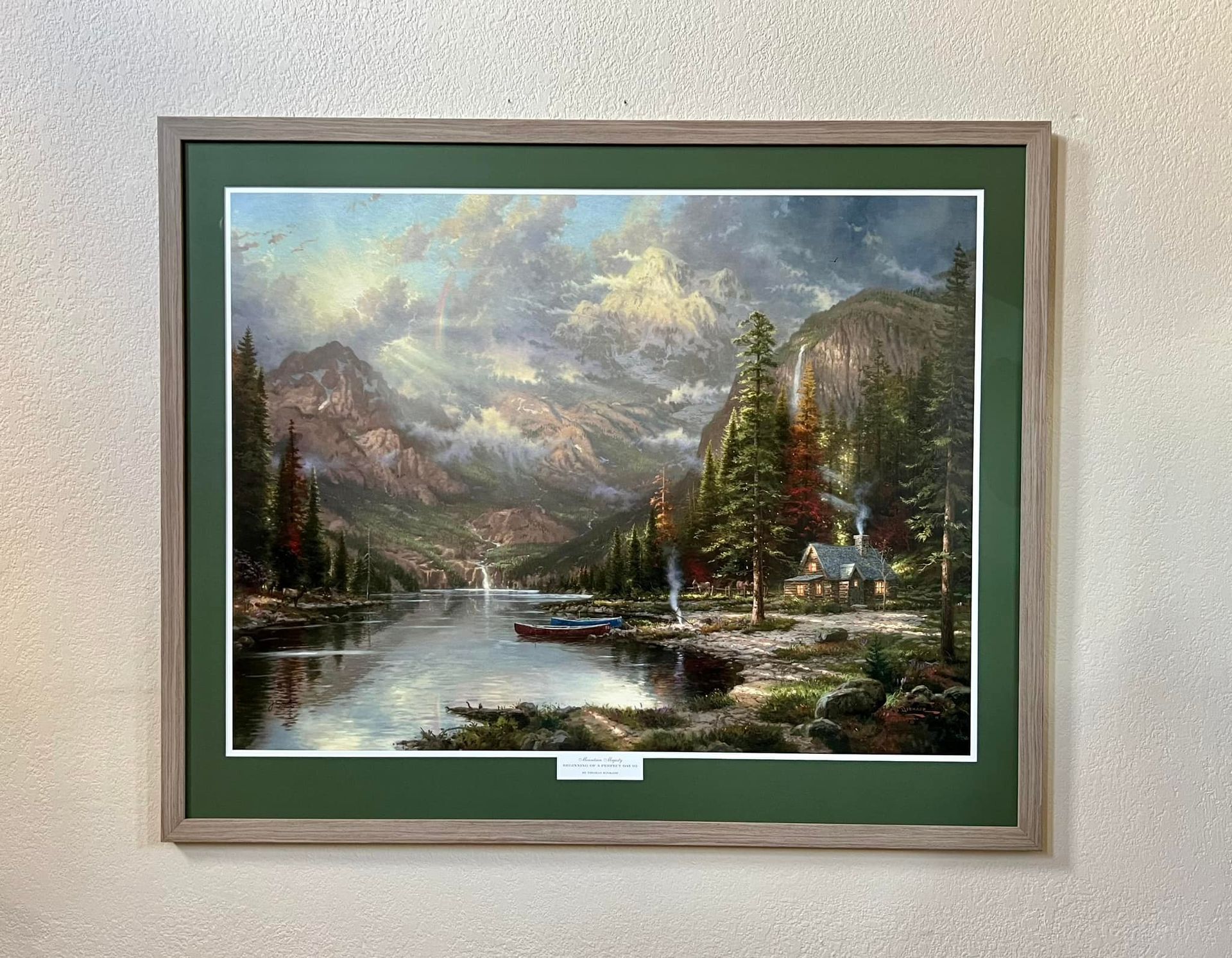 Framed landscape painting with mountains, lake, trees, boats, and a cabin; green mat and brown frame.