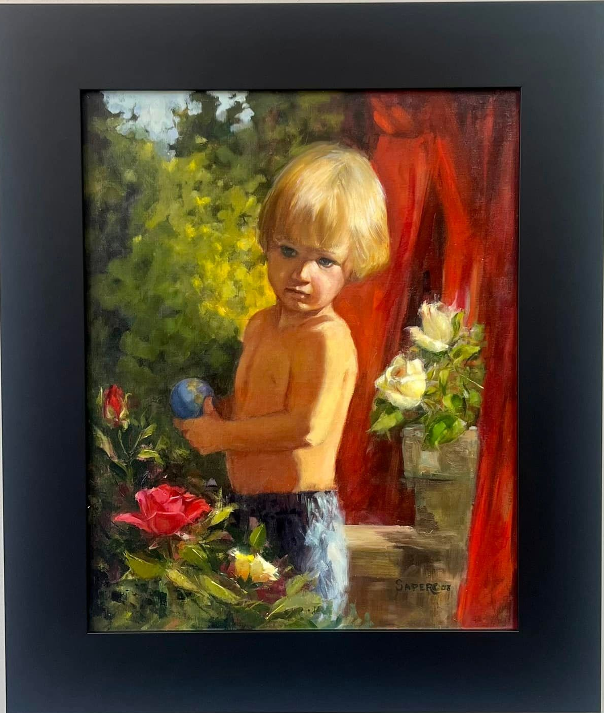 Blond child, shirtless, holding a blue ball, stands among roses and a red curtain.