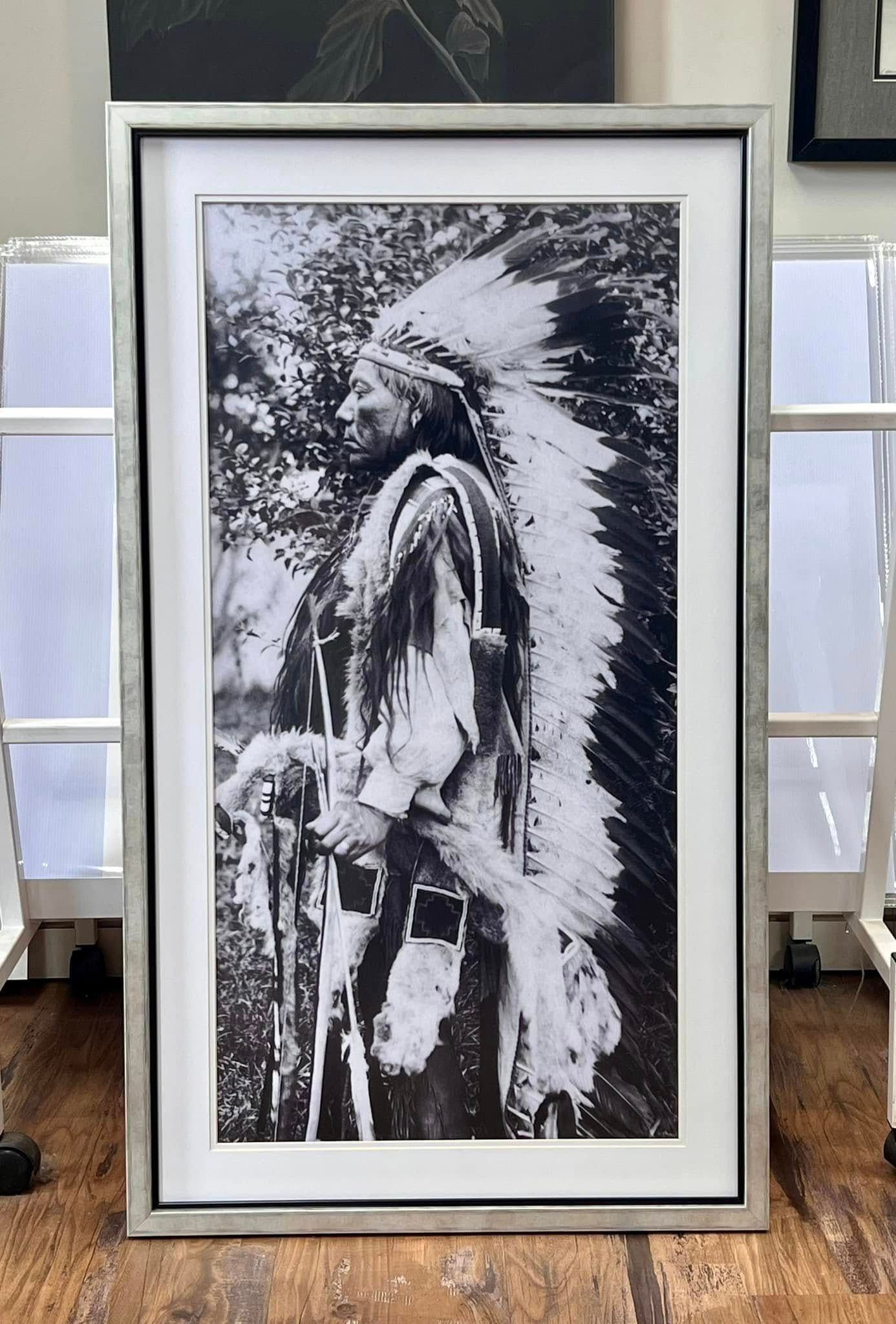 Black and white photo of a Native American man in profile wearing a feathered headdress and holding items, framed.