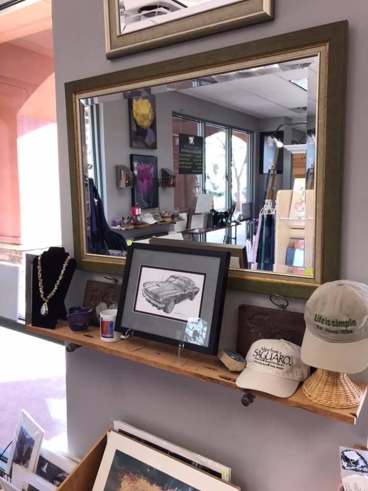 A store display with a mirror, framed artwork, jewelry, hats, and other merchandise on a wooden shelf against a gray wall.