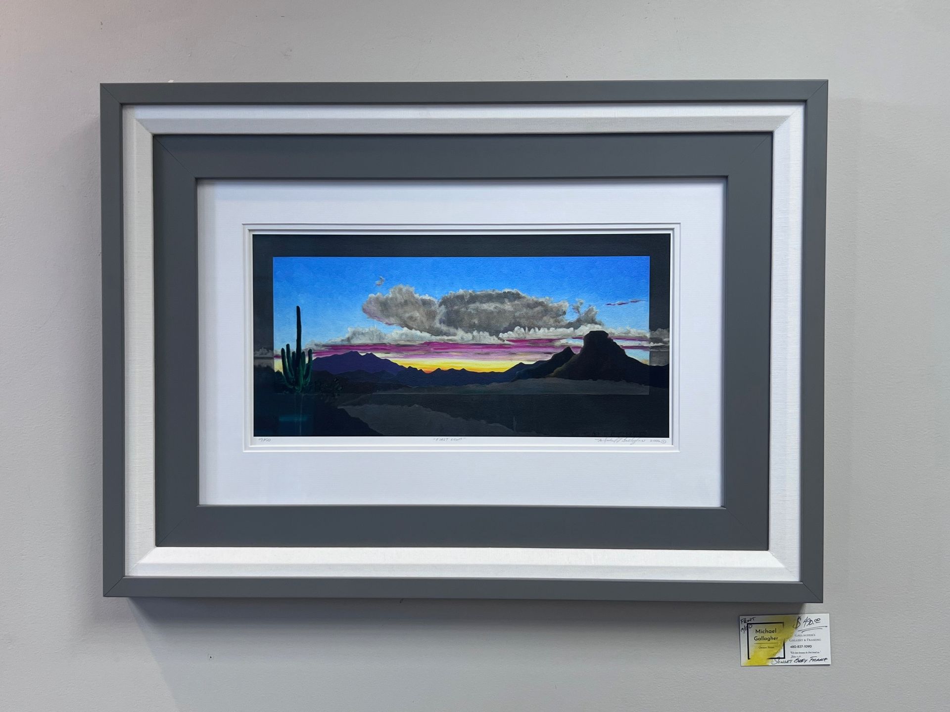 Framed artwork of a colorful sunset over a desert landscape, dark mountains, and a cloudy sky.