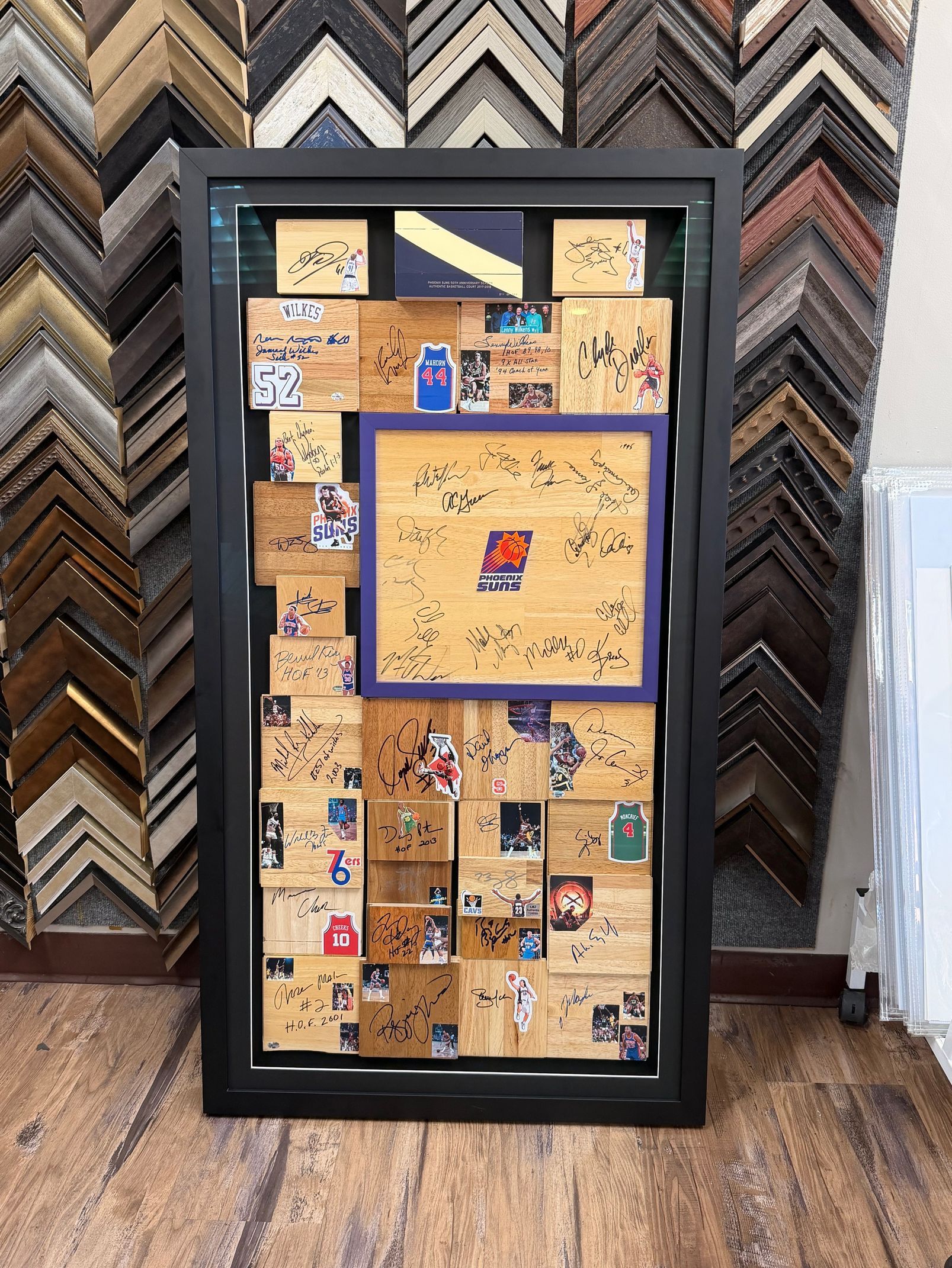 Black framed artwork with layered corkboard tiles, decorated with small images and stickers. Variety of frame samples in background.