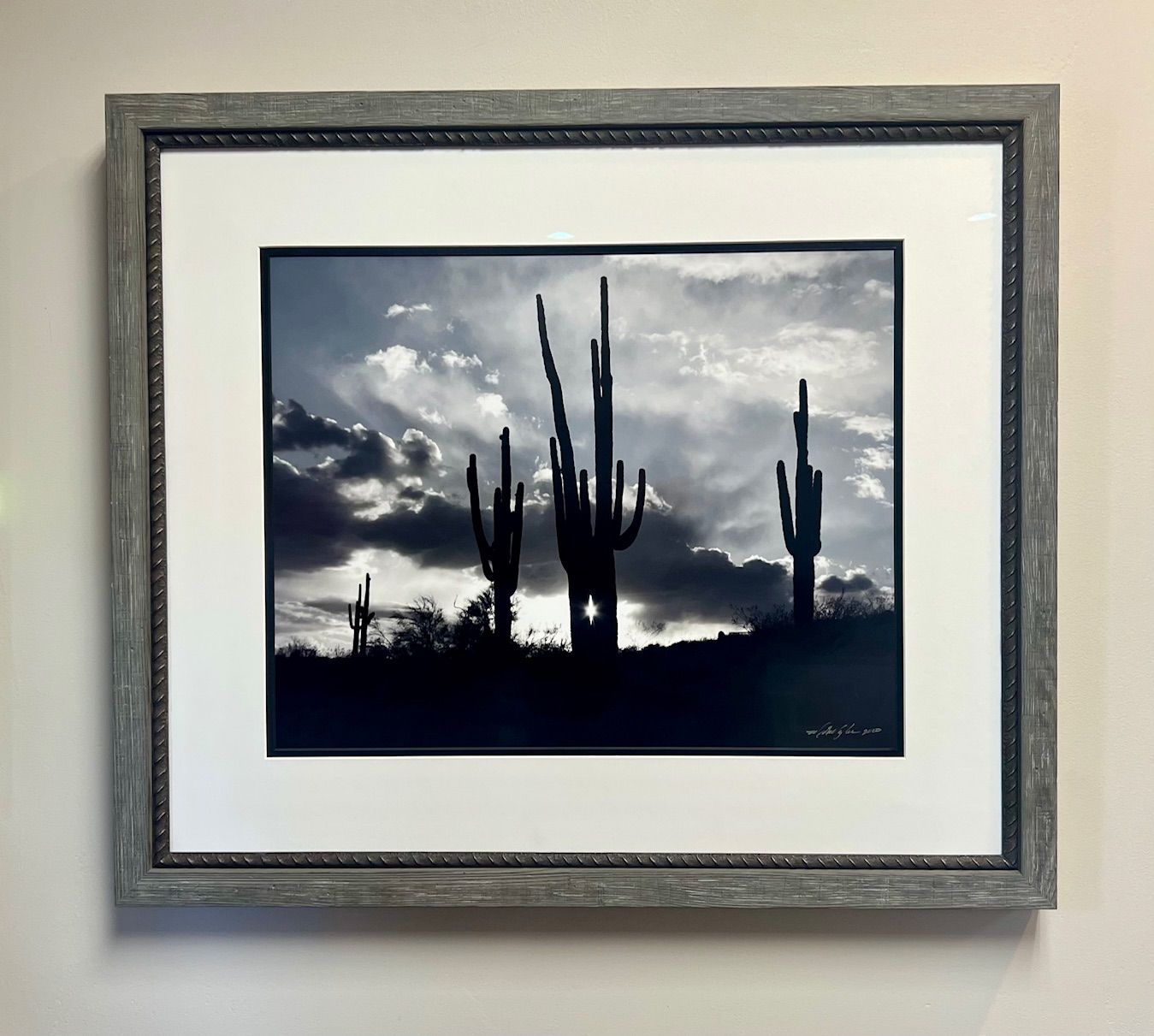 Colorful folk art landscape painting in a black frame, depicting a village, trees, volcano, and water.