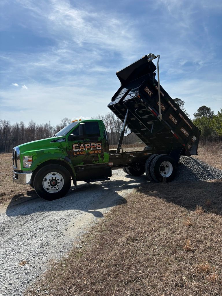 Green dump truck with raised bed on gravel, loading or unloading debris outdoors under a blue sky