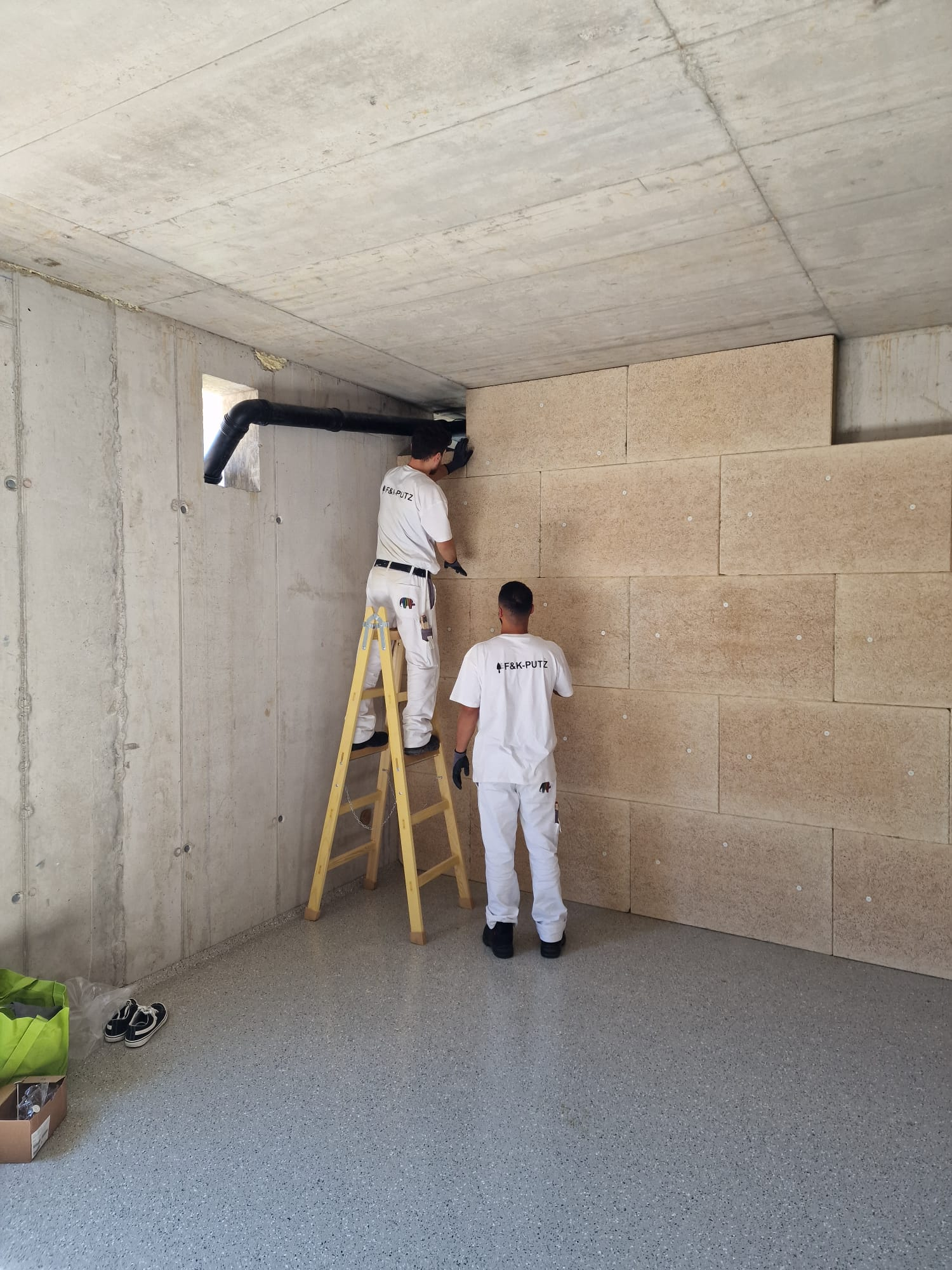 Zwei Männer arbeiten mit einer Leiter an einer Wand in einem Raum.