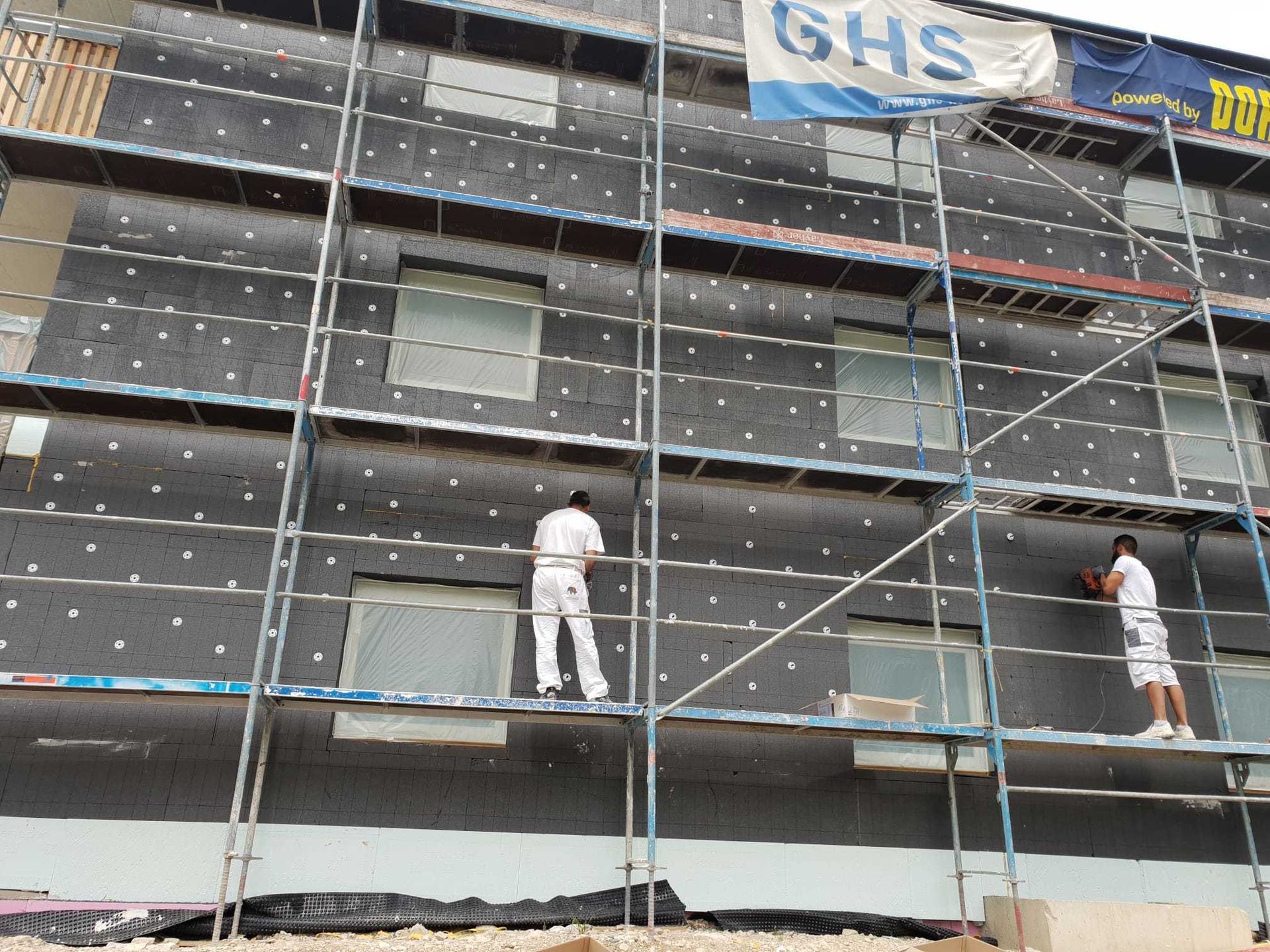 Zwei Männer stehen auf einem Gerüst an der Seite eines Gebäudes unter einem GHS-Banner
