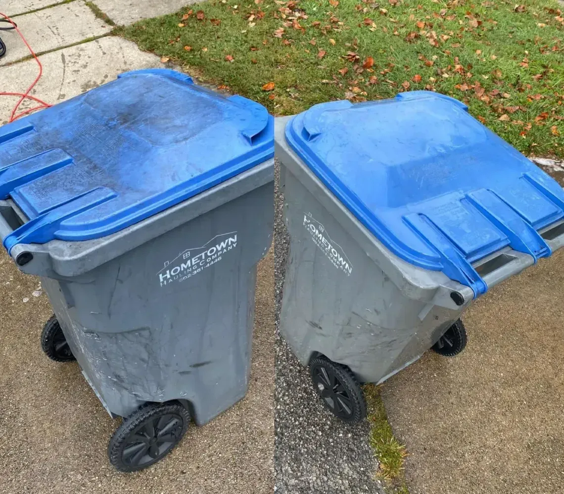 Two grey plastic waste bins with blue lids sit side-by-side on a concrete driveway.