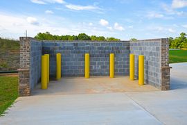An outdoor waste enclosure with gray concrete block walls and six vertical yellow protective bollards on a concrete pad.