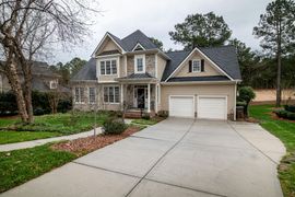 A beige two-story house with a stone facade, dark roof, and a concrete driveway in a suburban neighborhood.