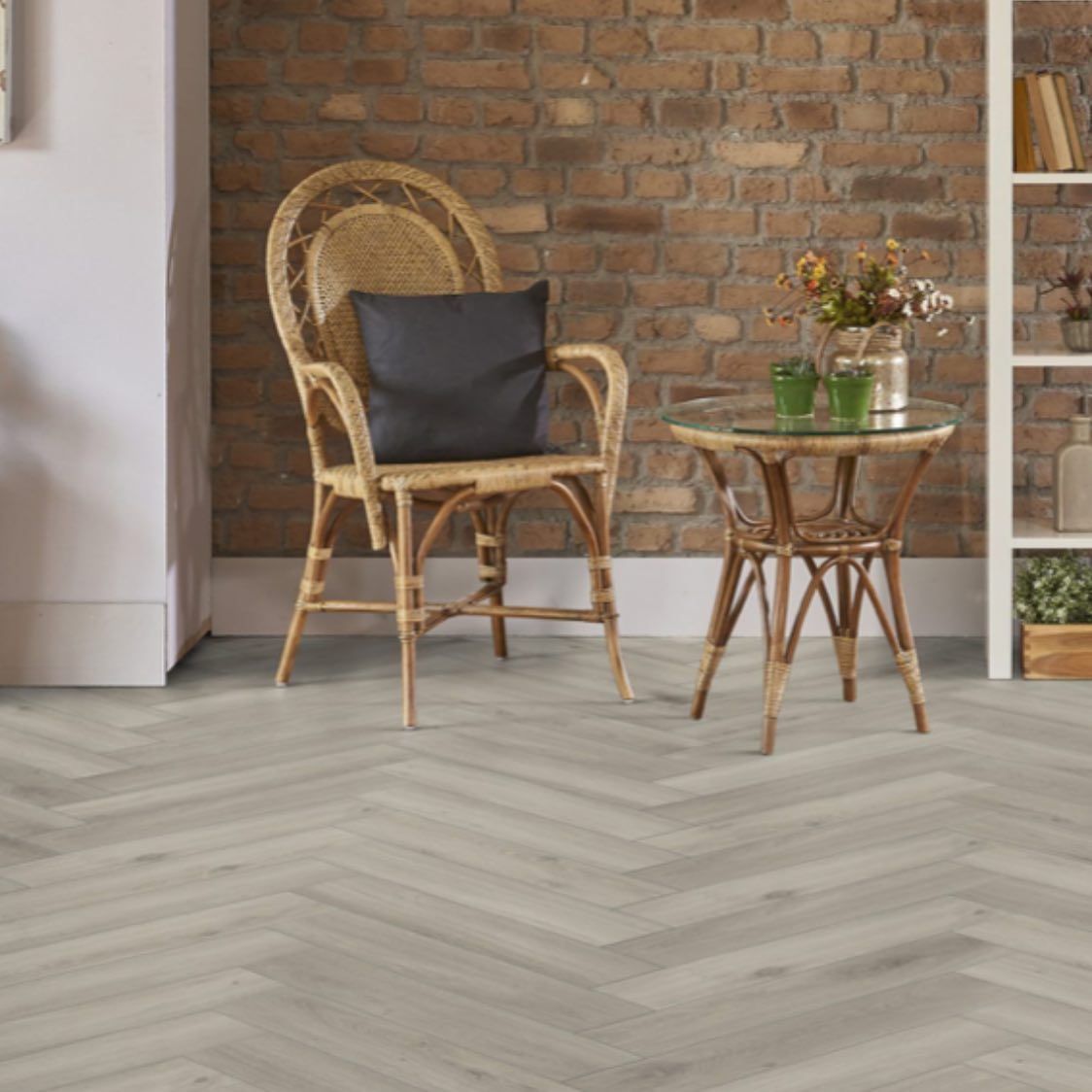 Herringbone pattern wood flooring in a room with a wicker chair, table, and brick wall.