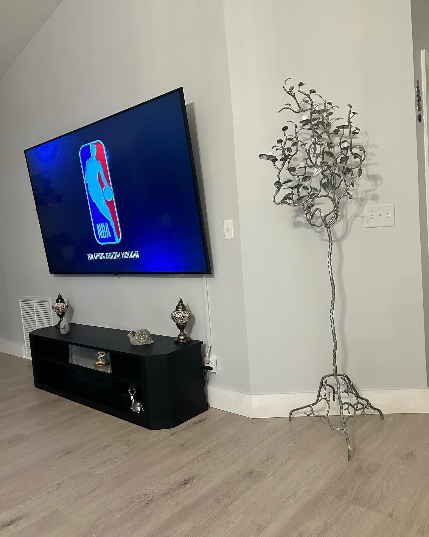 A modern living room with a TV mounted on a gray wall, black entertainment center, and decorative metal tree.
