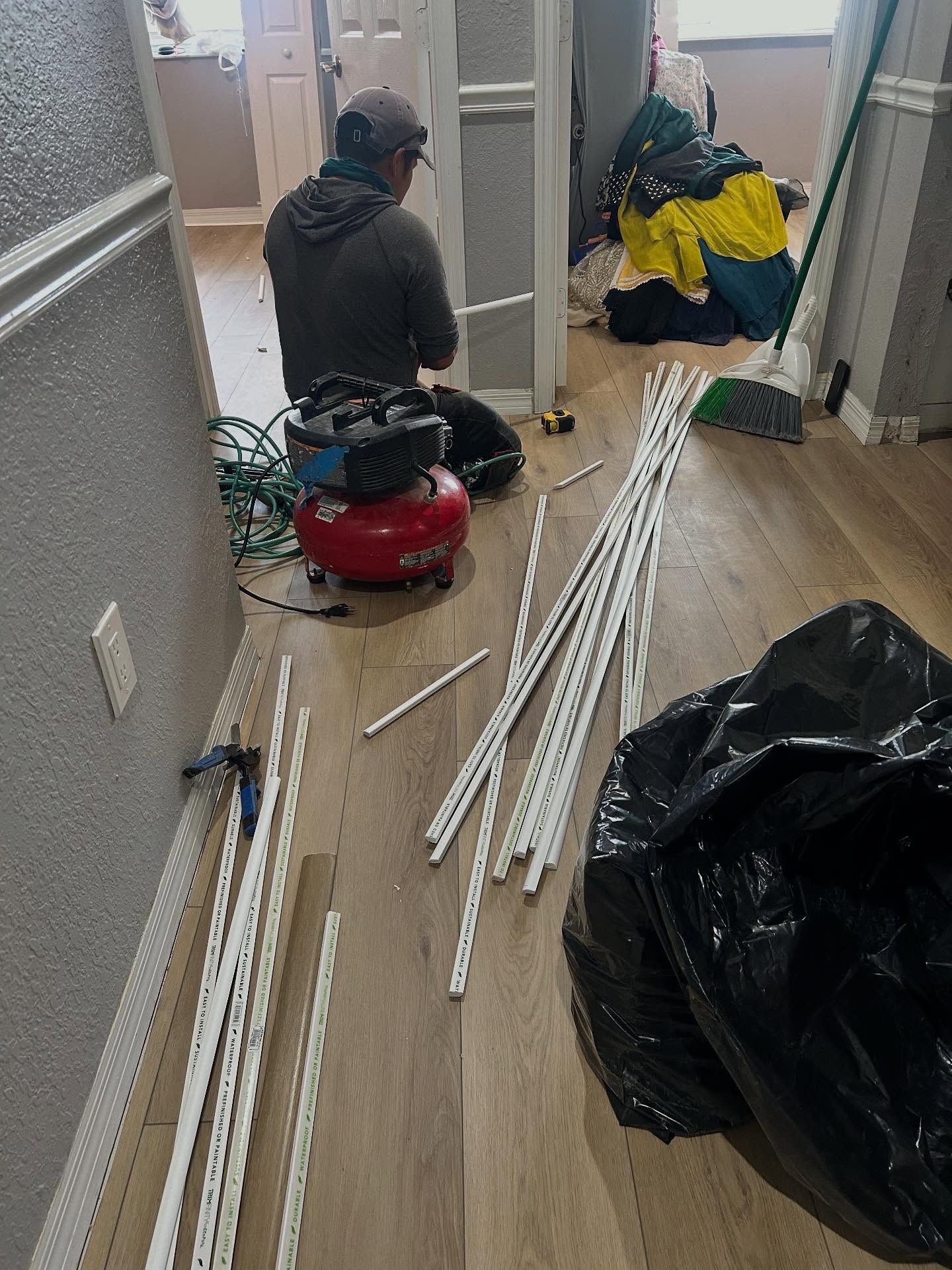 Person installing trim, red air compressor and tools on floor, white trim pieces and black trash bag in room.