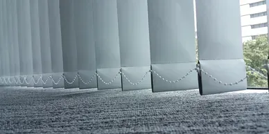 White vertical blinds hang in front of a window, with a carpeted floor in the foreground.