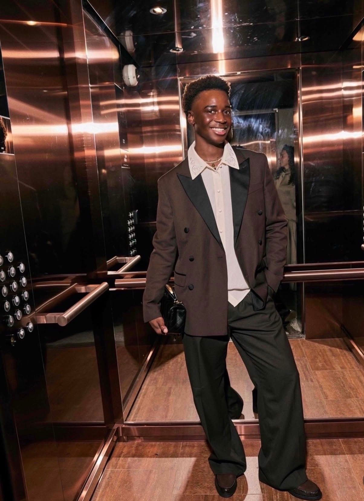 Een persoon in een bruine blazer staat in een lift met spiegelwanden, glimlachend met één hand in zijn zak.