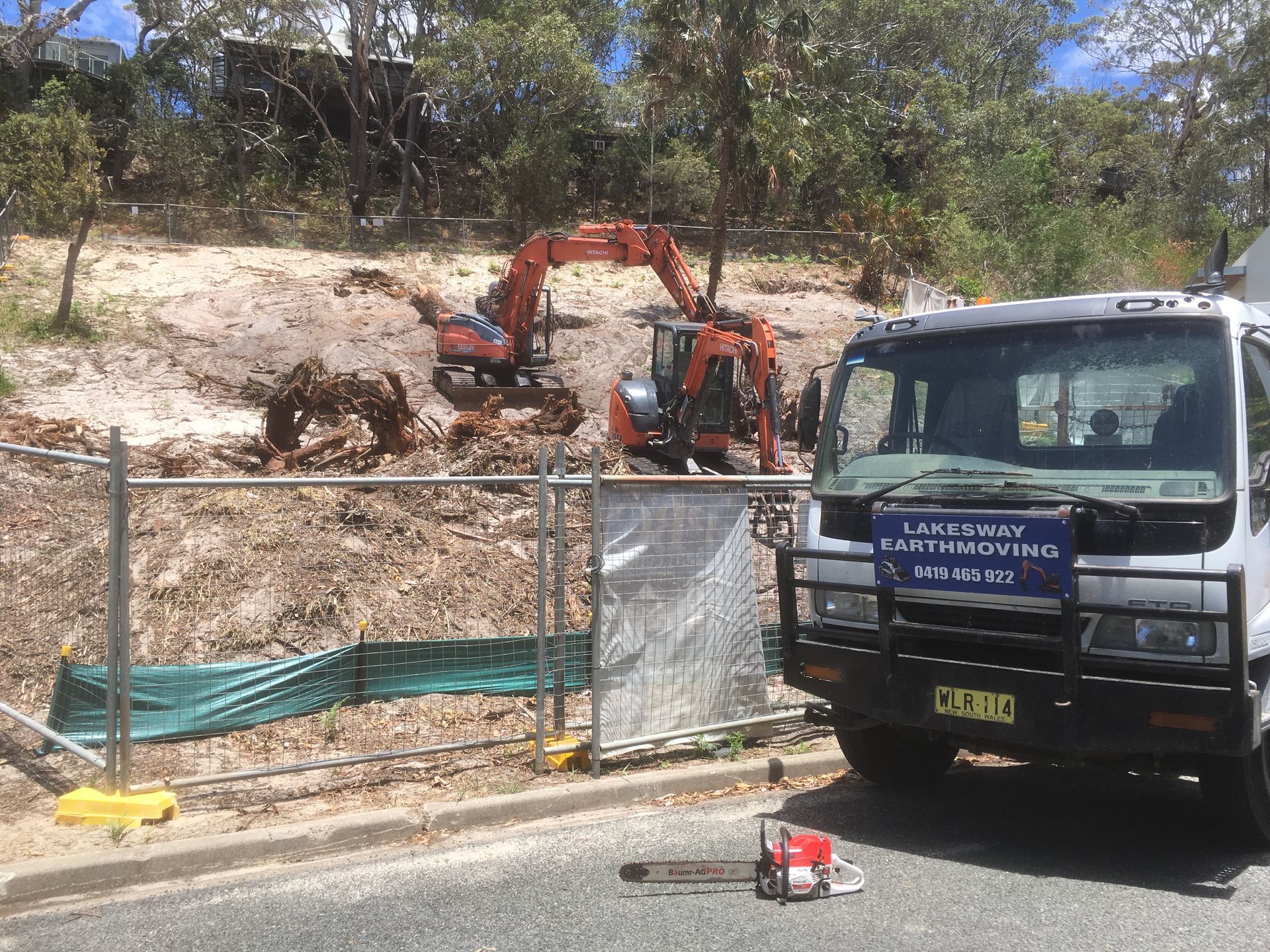 Construction site with excavator, truck, and chainsaw. Earthmoving equipment working on a hillside, fenced off.