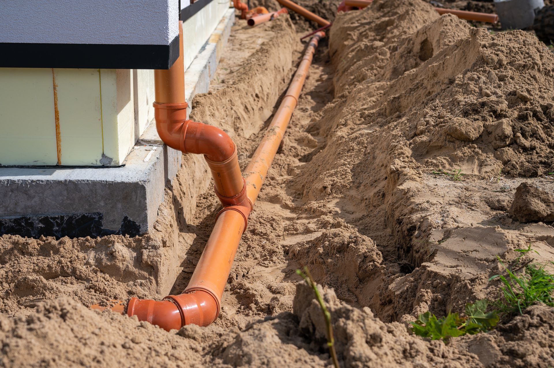 Orange drainage pipes installed in a trench next to a building foundation in sand.