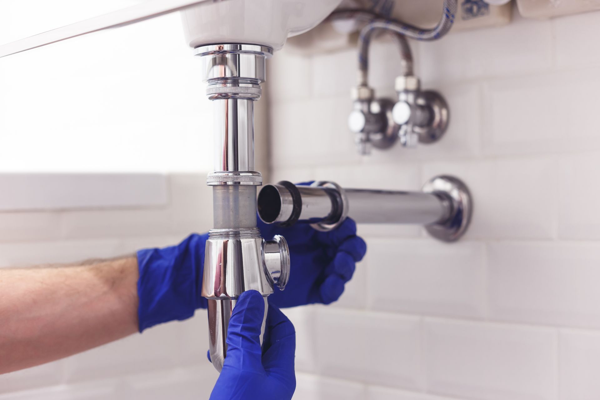 Hands in blue gloves assembling plumbing under a white sink in a tiled bathroom.