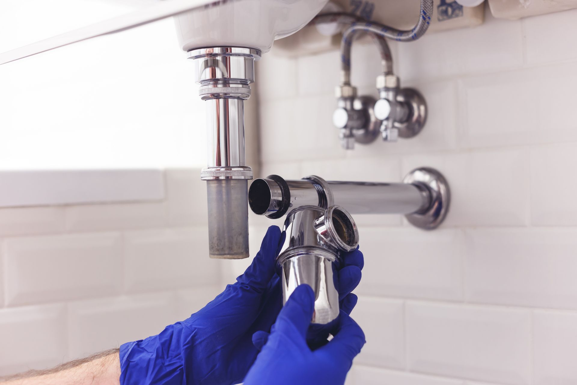 Hands in blue gloves assembling a chrome sink trap under a white sink in a tiled bathroom.