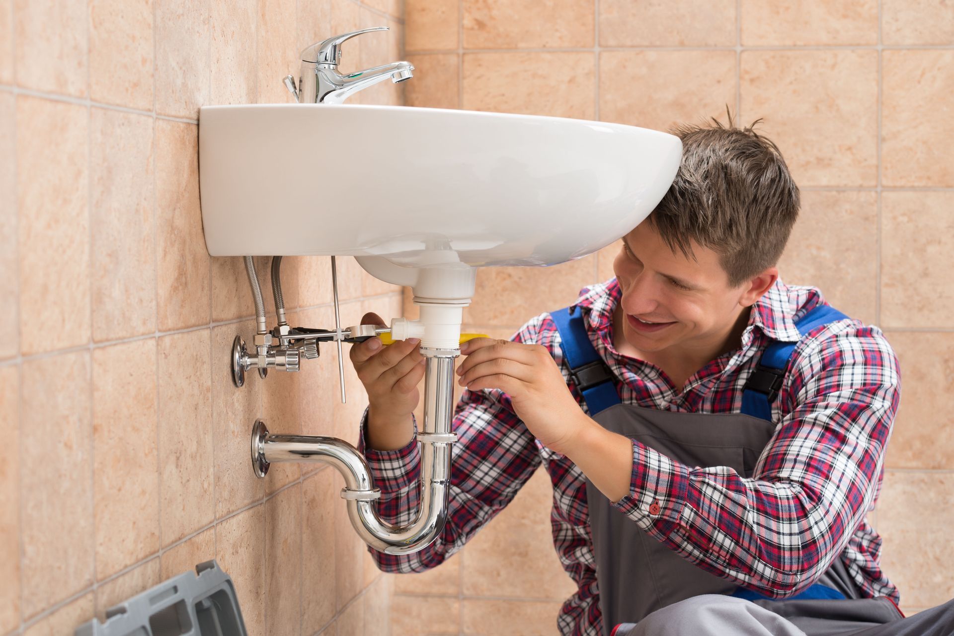 Plumber smiling while working on a bathroom sink; beige tiles in background.