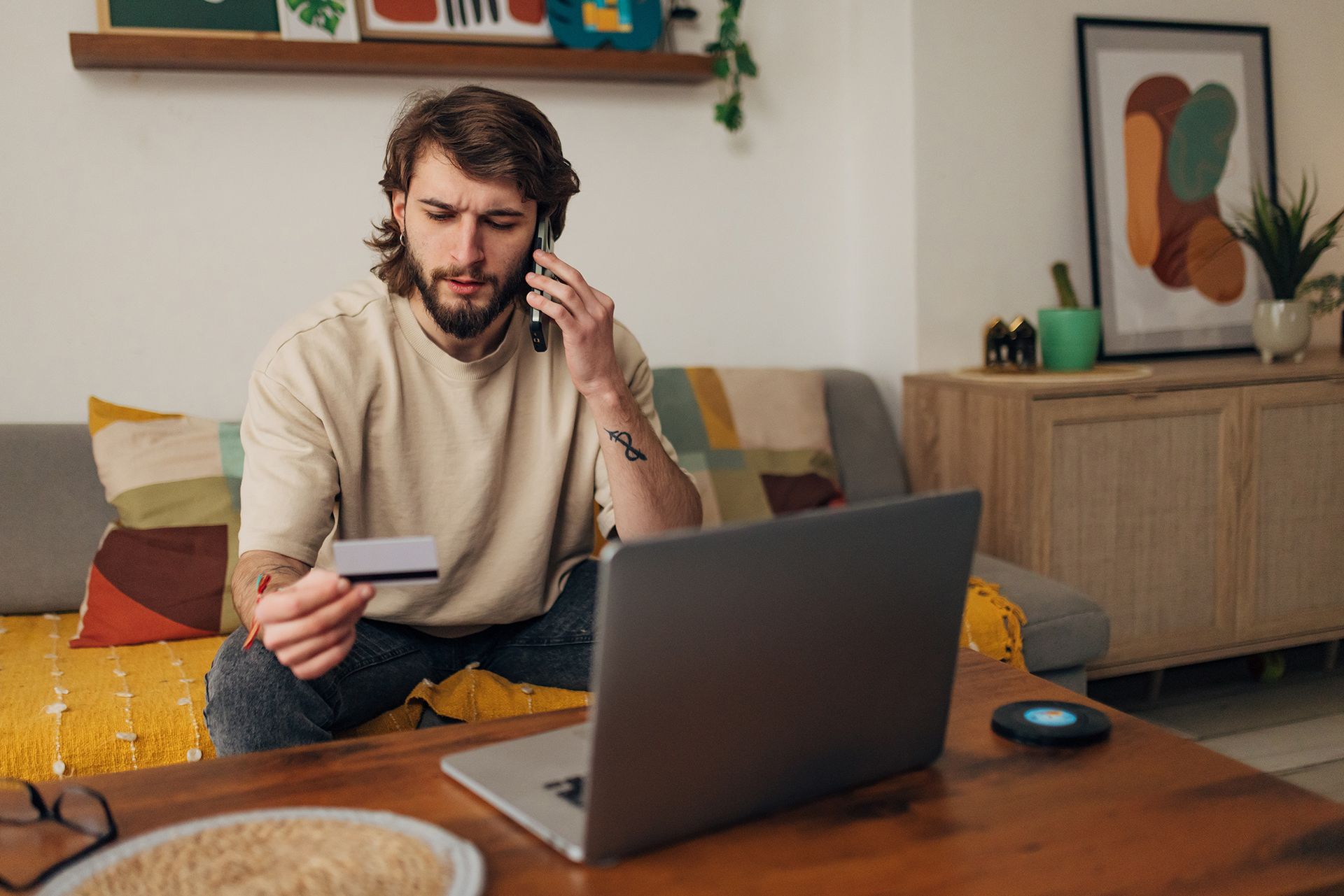 A man is sitting on a couch holding a credit card and talking on a cell phone.