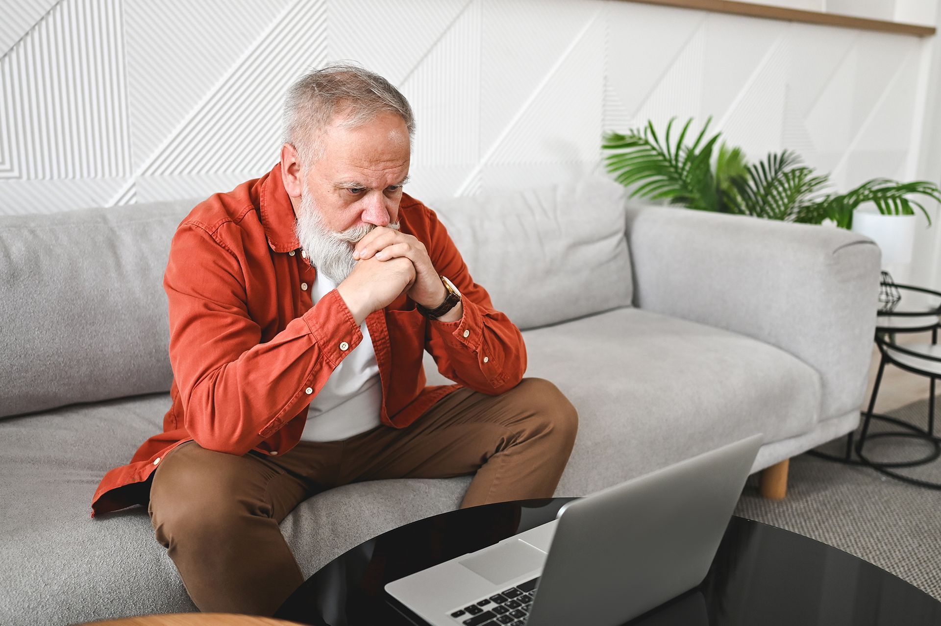 An older man is sitting on a couch looking at a laptop computer.