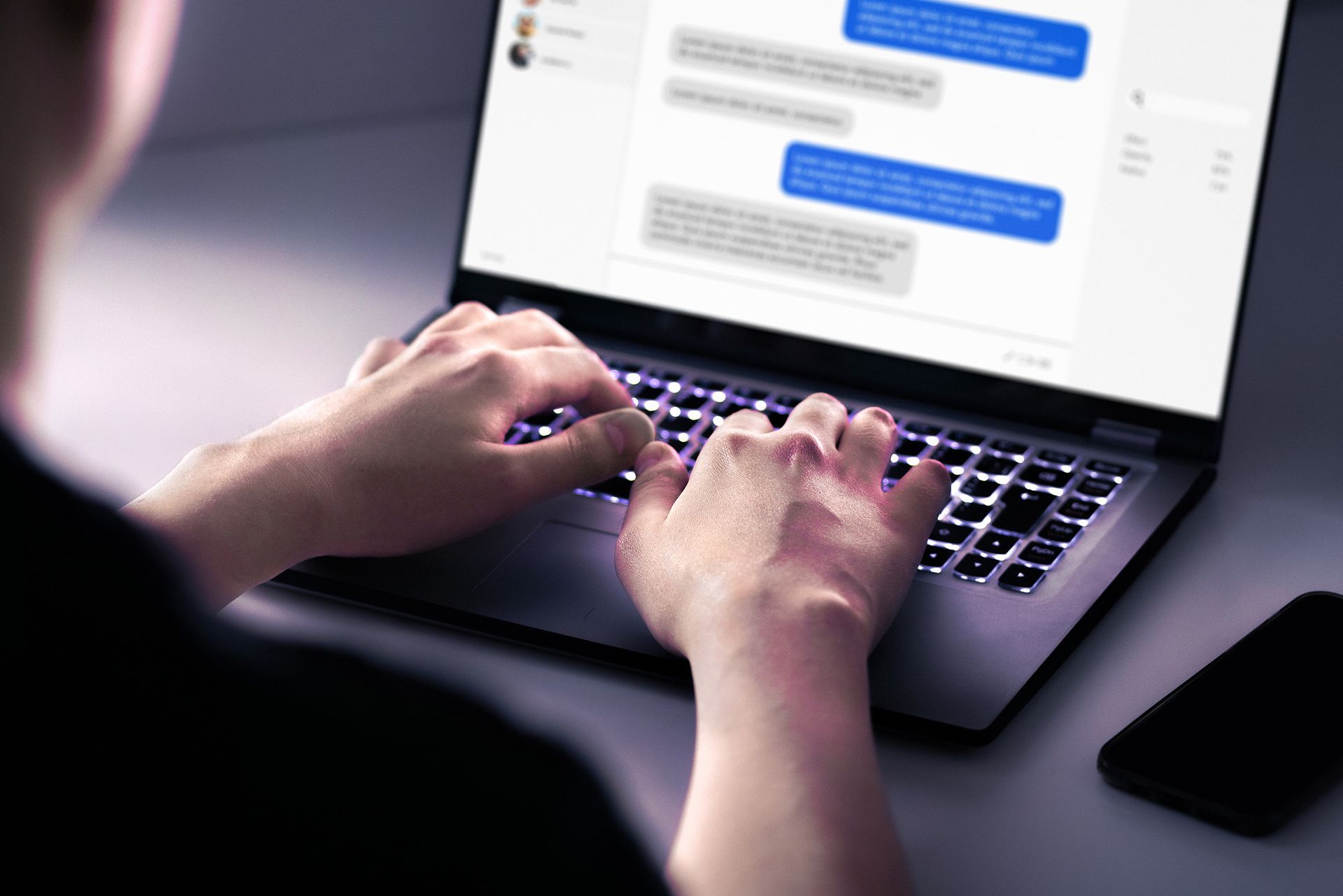 A person is typing on a laptop with a phone in the background.