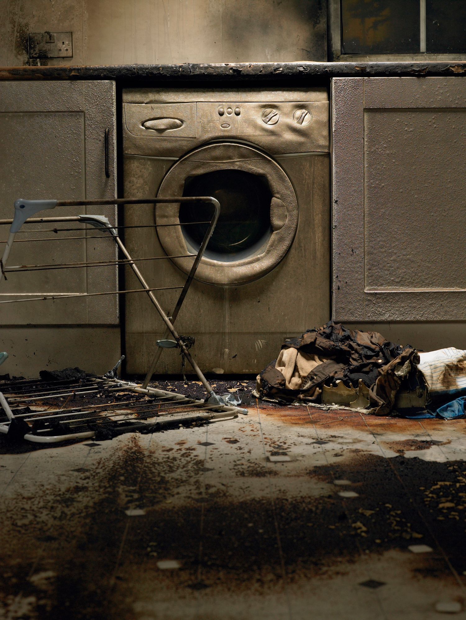 Burnt washing machine in a kitchen, charred cabinets and floor, drying rack, damaged clothes.