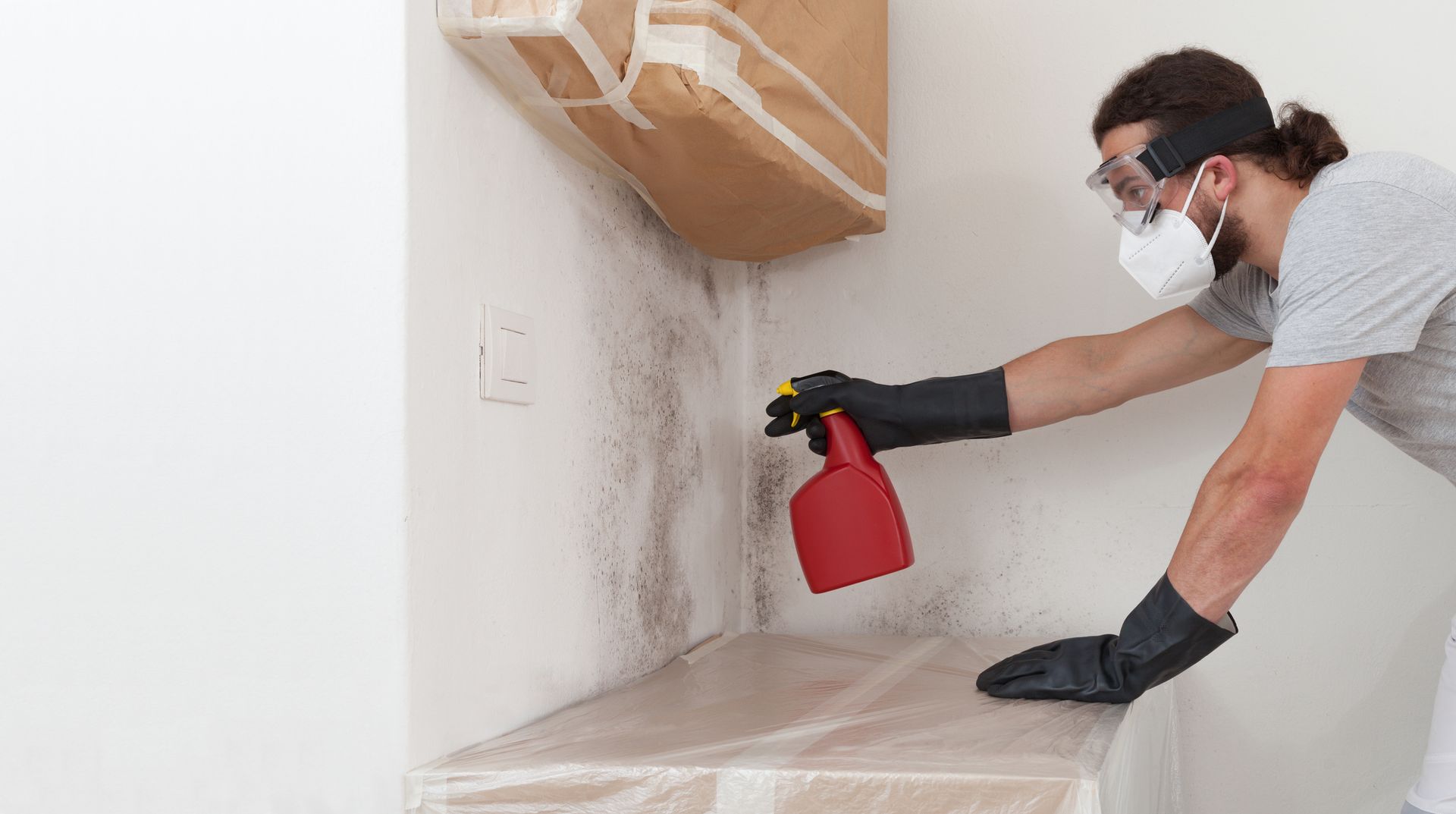 Man spraying mold remover on wall wearing gloves and respirator during mold remediation.