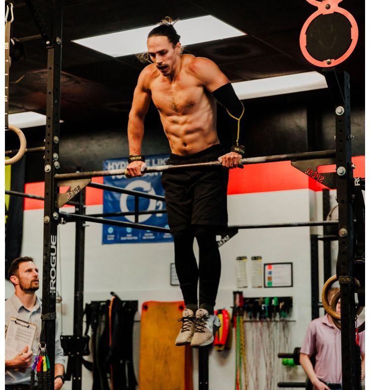 A person performs a bar dip in a Tallahassee gym, with a coach observing and a person standing in the background.