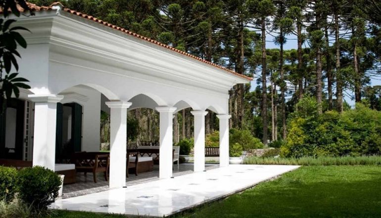 Uma casa branca com colunas e arcos em frente a uma floresta