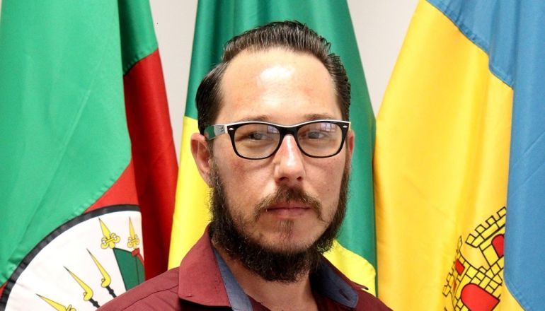 Um homem de óculos e barba está parado em frente a bandeiras.