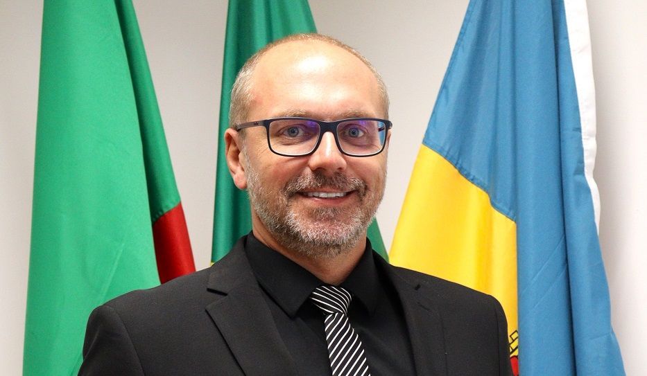 Um homem de terno e gravata está diante de três bandeiras.