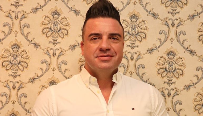 Um homem de camisa branca está parado em frente a uma parede com papel de parede estampado.