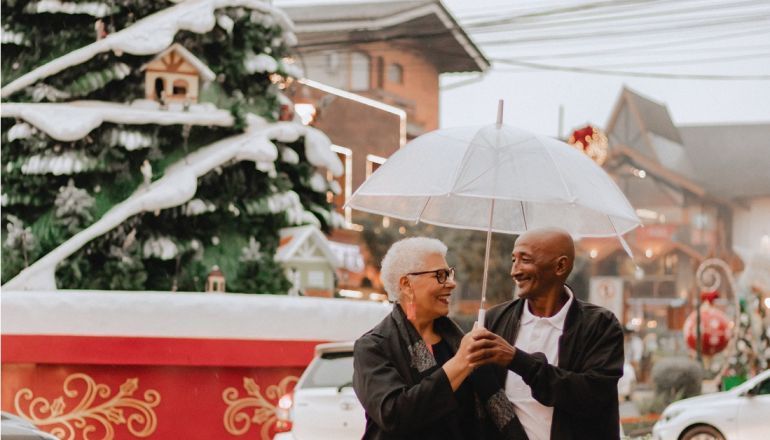 Um casal de idosos está segurando um guarda-chuva na chuva em frente a uma árvore de natal.