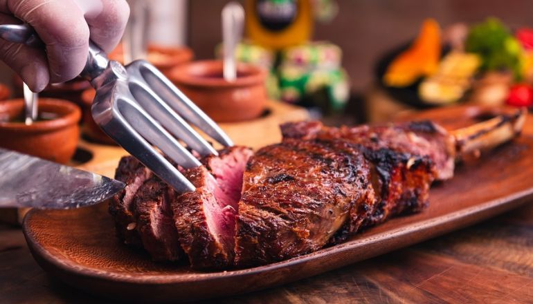 Uma pessoa está cortando um bife com um garfo em uma tábua de madeira.