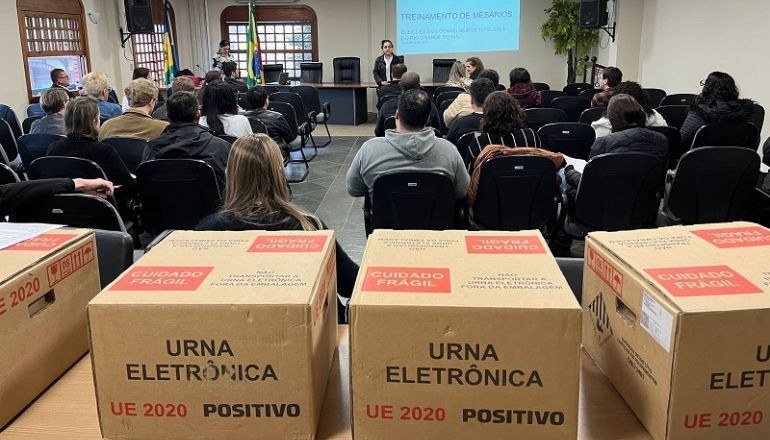 Um grupo de pessoas está sentado em uma sala com caixas que dizem urna eletrônica.
