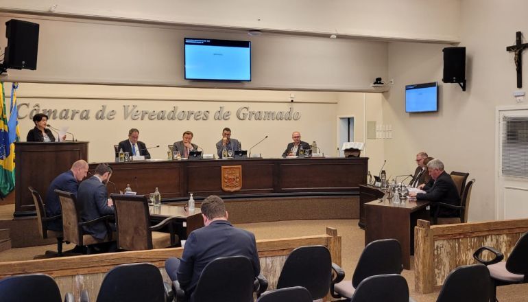 Um grupo de pessoas está sentado em uma sala com uma placa que diz camara de vereadores de gramado